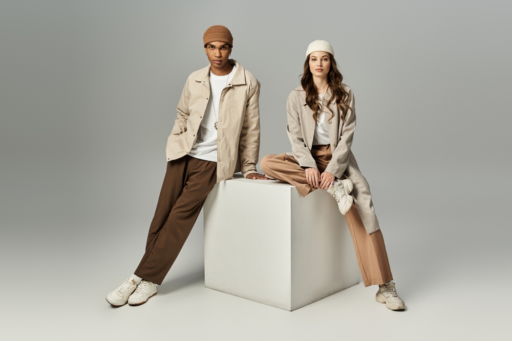 A Man and woman layering a lightweight sweater over a white shirt with a stylish coat and trench coat for a chic seasonal look.
