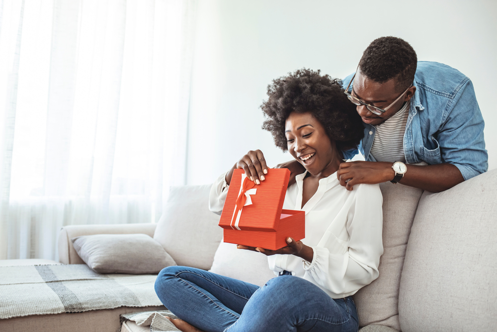 A joyful African American couple, with the male presenting a red gift box to the female, who smiles warmly in appreciation