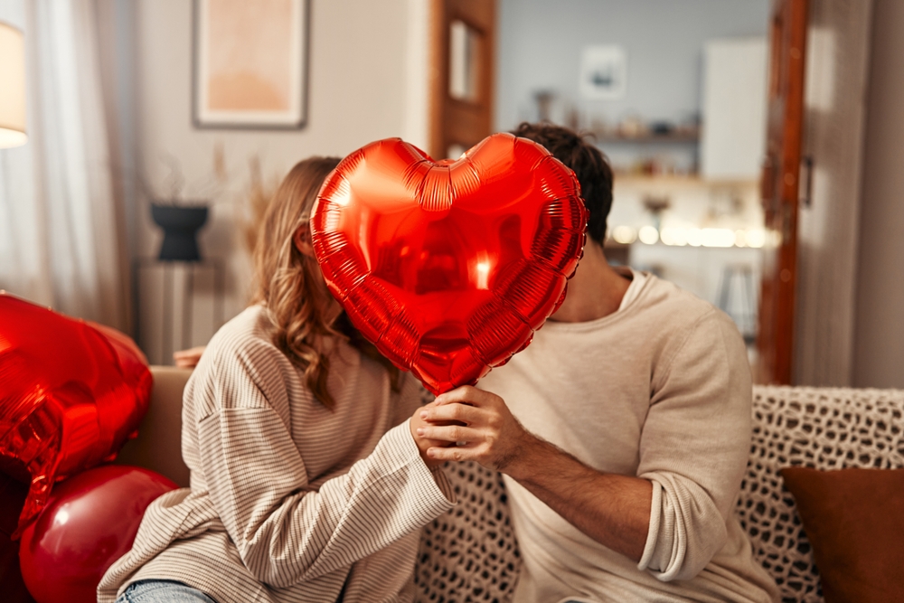 Man and woman holding a heart-shaped love balloon, smiling at each other in a romantic setting.
